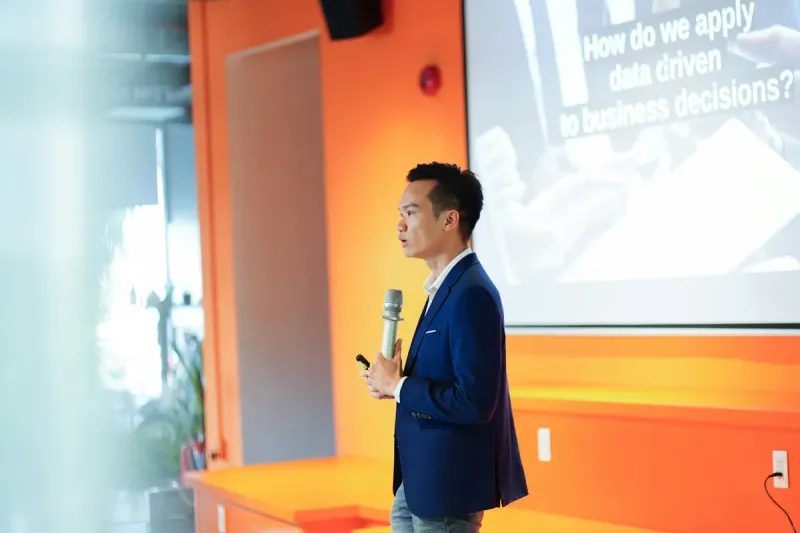 A man in a blazer speaks into a microphone on an orange-lit stage, with a slide reading "How do we apply data driven to business decisions?" behind him.