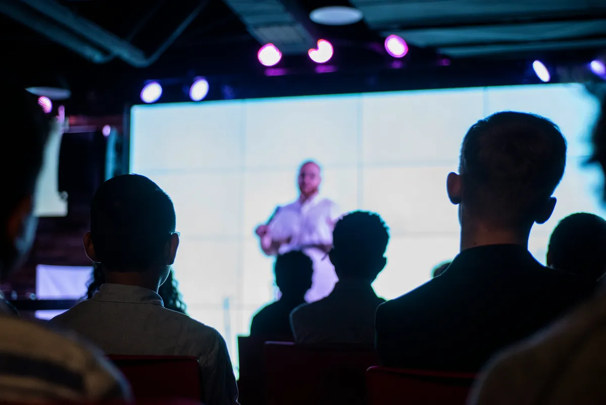An out-of-focus audience watches a presenter speaking in front of a large screen.