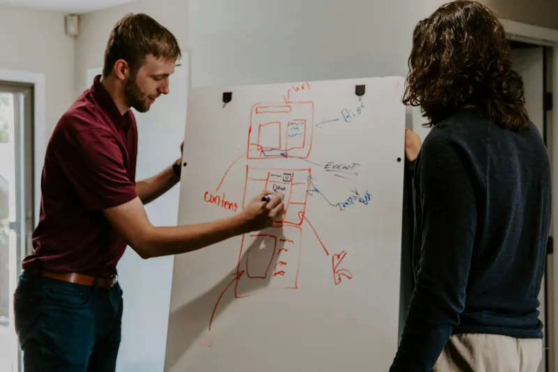 Two people at a whiteboard, one sketching a diagram while the other looks on.
