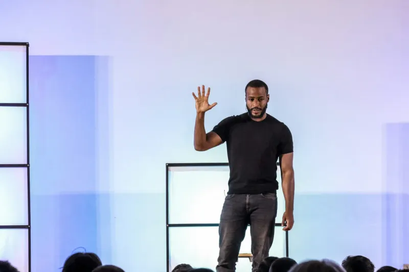 A presenter on stage raises one hand with fingers spread, addressing an audience.