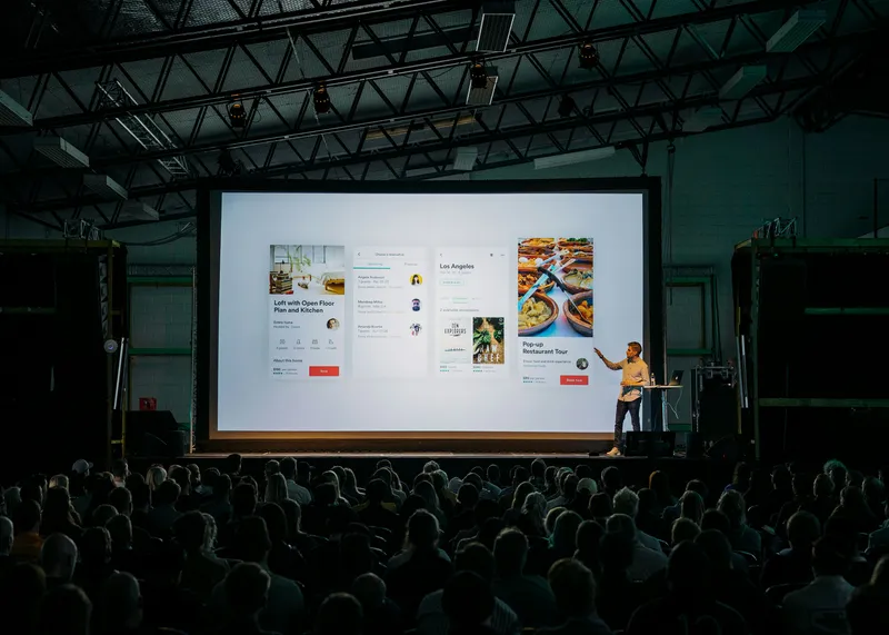 A presenter addresses a large seated audience in an industrial-style venue, standing beside a big screen showing app design slides.