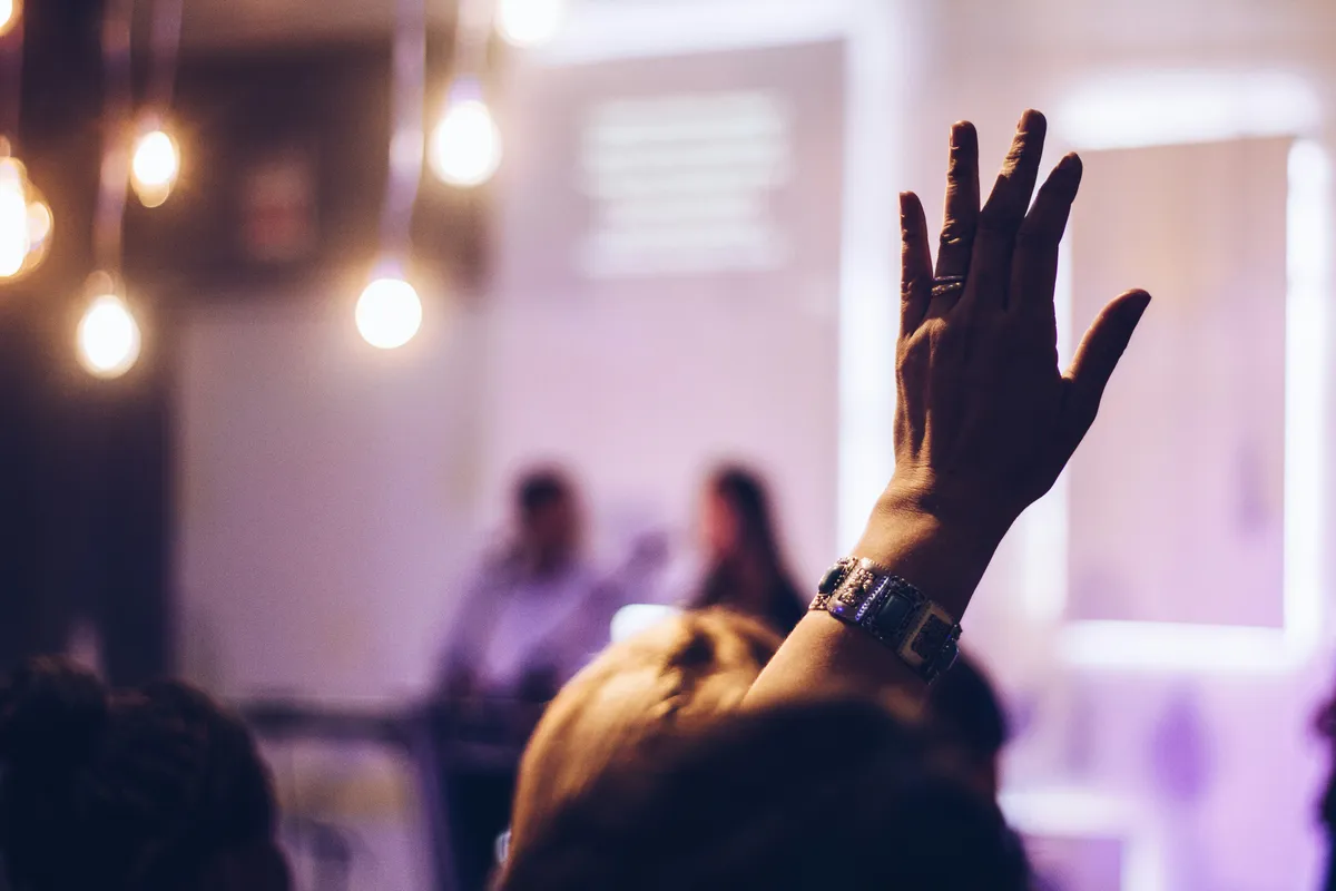 A close-up of a hand raised in an audience, with blurred string lights in the background.