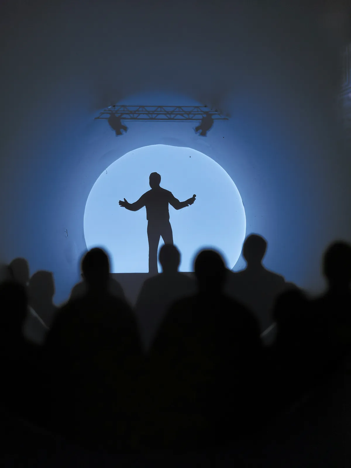 A silhouetted speaker stands with arms outstretched in a blue spotlight, seen from behind a seated audience.