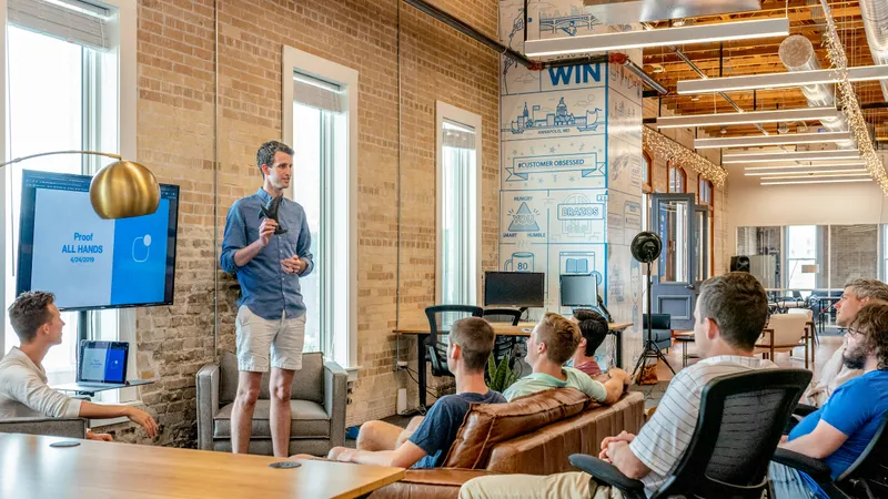 A man presents to colleagues seated around a couch in a modern office, with a screen and wall graphics reading "WIN" behind them.